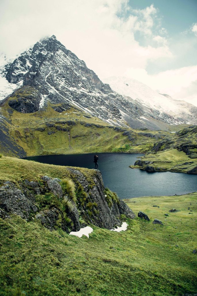 A hiker stands on a cliff overlooking a serene mountain lake surrounded by lush greenery and snow-capped peaks.